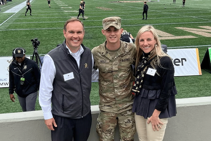 A U.S. Army cadet poses with two smiling adults on a football field sideline before a game.