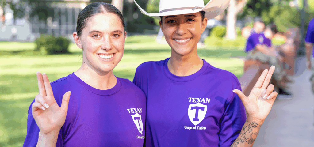 Two smiling cadets in purple Corps of Cadets shirts pose outdoors, making the States Up hand sign.