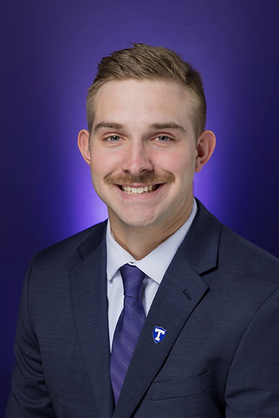 Man in dark suit and purple tie smiles in front of a purple background with Tarleton State University pin.