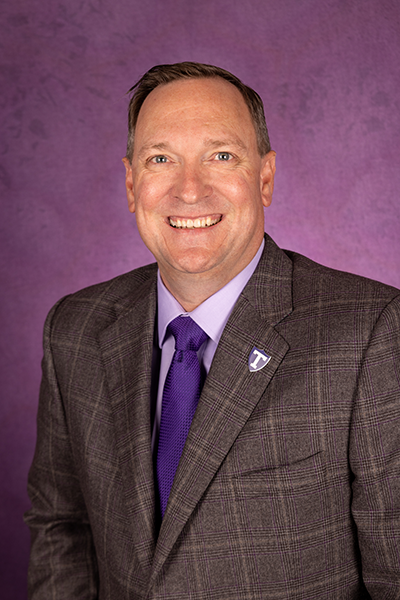 Man in a gray plaid suit with a purple tie smiling in front of a solid purple background.