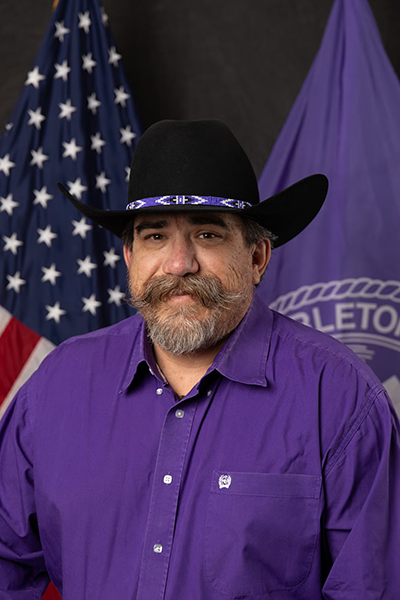 Man wearing a black cowboy hat and purple shirt poses in front of U.S. and Tarleton State University flags.