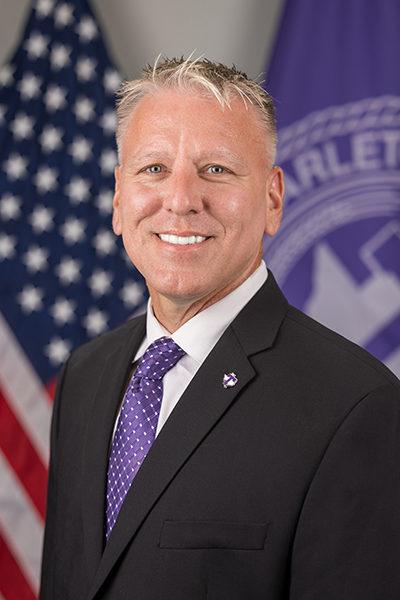Main in a dark suit and purple tie smiling in front of the American flag and a purple Tarleton State University flag.