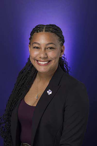Woman in dark blazer and maroon top smiles in front of a purple background with Tarleton State University pin.