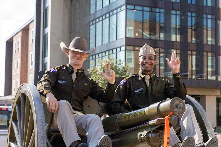 Two Tarleton cadets in uniform smile and pose with the Tarleton hand sign while sitting on a cannon in front of campus buildings.