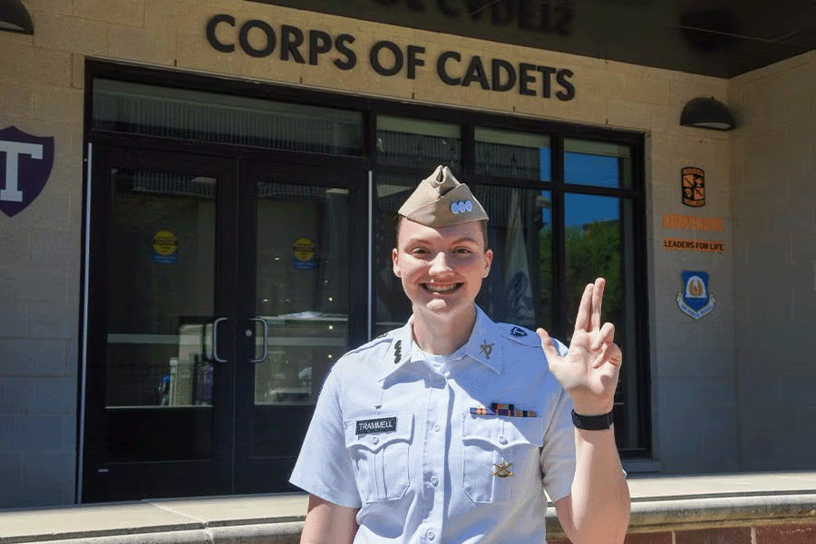 Tarleton cadet in uniform smiles and gives the Tarleton hand sign in front of the Corps of Cadets building.