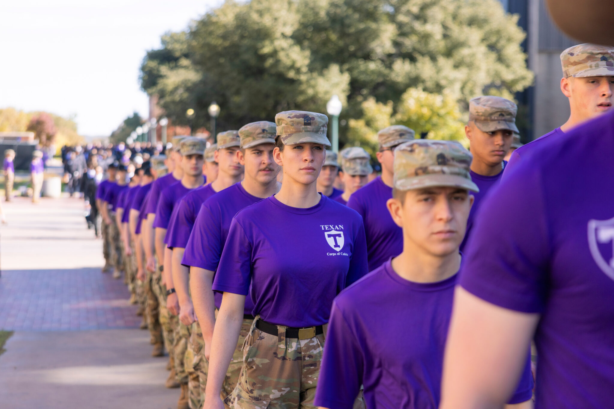 Cadets in purple shirts and camo caps march in a long, orderly formation down a campus walkway.