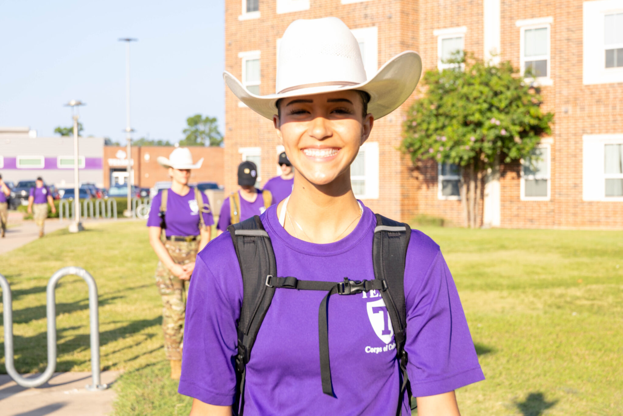Smiling cadet in a white cowboy hat and purple Tarleton Corps shirt stands outdoors with other cadets.